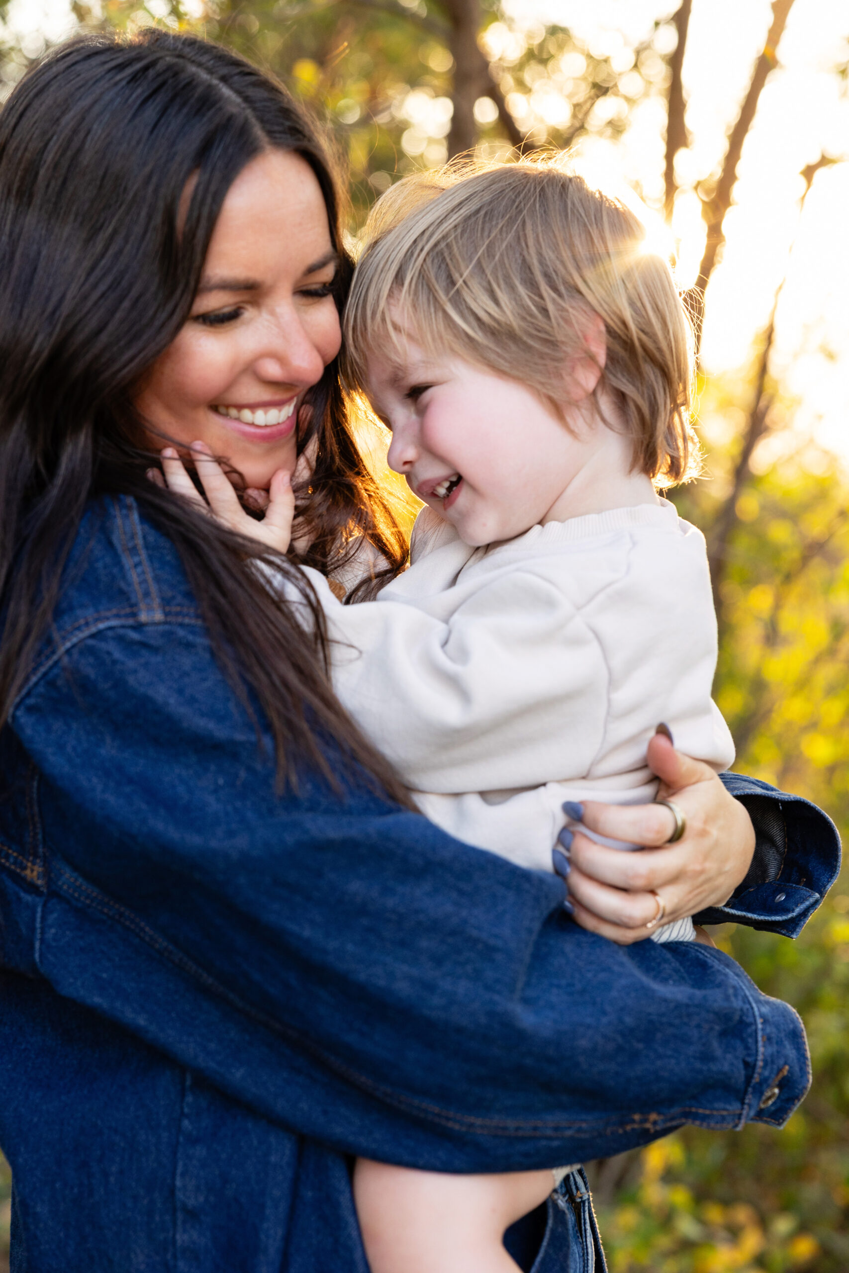 Woman in a denim jacket holding a young child outdoors at sunset, both smiling closely in warm light.