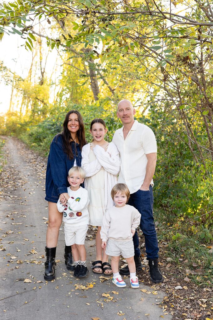 Family of five standing on a wooded path surrounded by golden fall leaves, smiling for a group portrait.