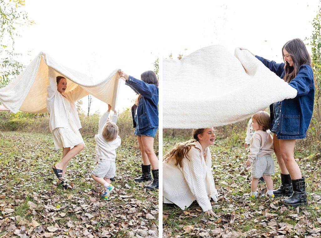 Family lifting a large cream-colored blanket over young kids in a clearing, surrounded by fall leaves and trees.