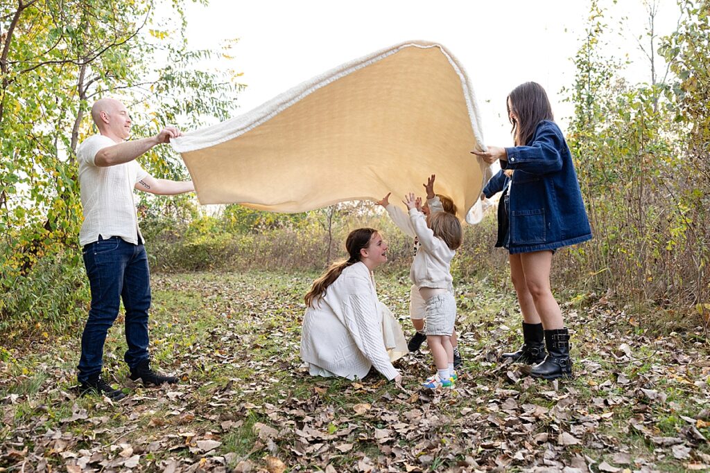 Parents and children playing with a large blanket in a wooded area, with dry leaves scattered on the ground.