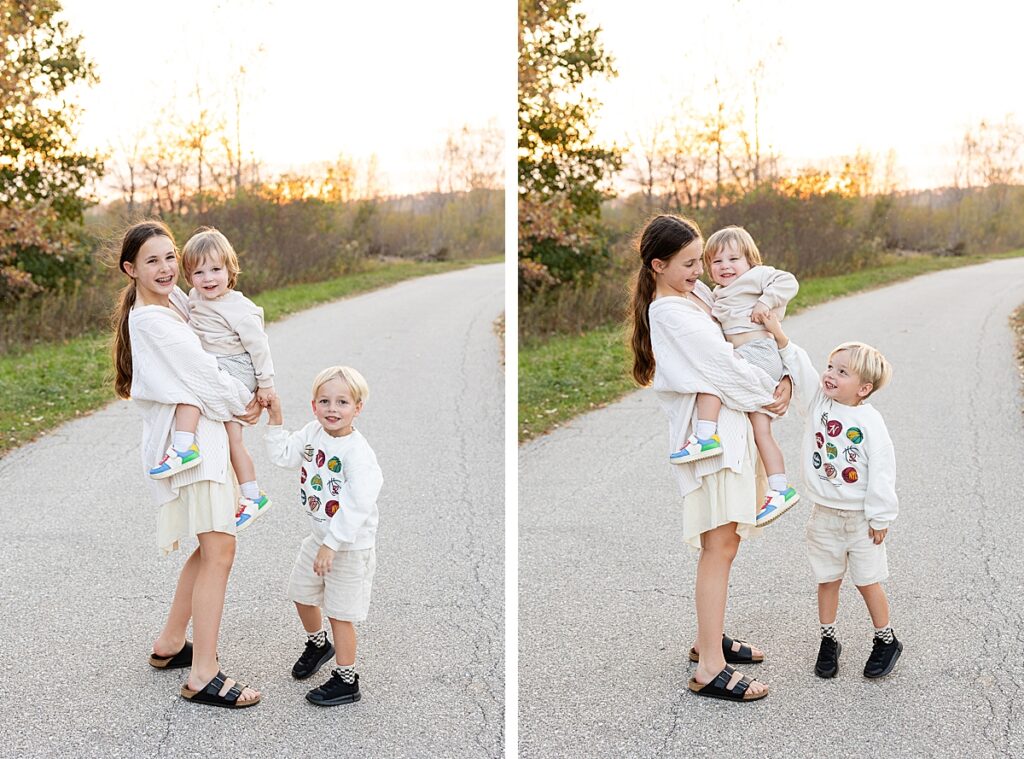 Young girl holding her toddler sibling with their brother beside them, smiling on a quiet path at sunset.