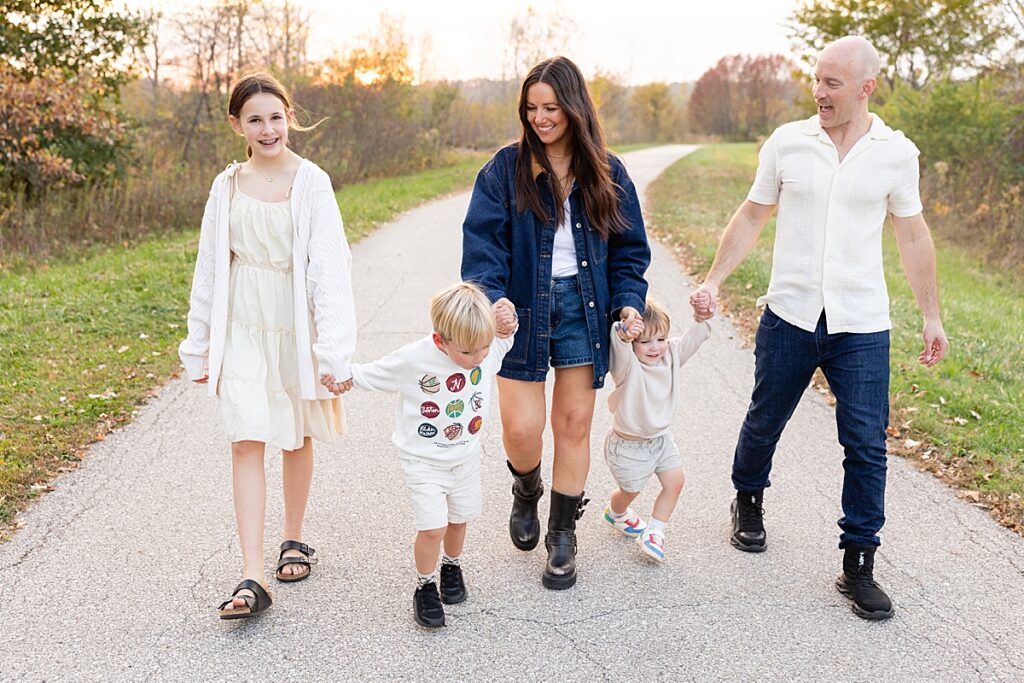 Family of five smiling and walking on a paved path at sunset, holding hands and playing along the way.