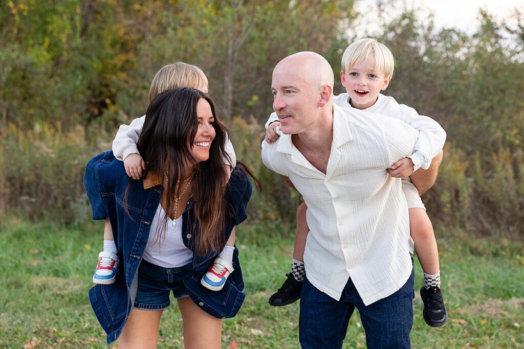 Parents giving piggyback rides to their two young sons in a grassy field, smiling and looking at each other.