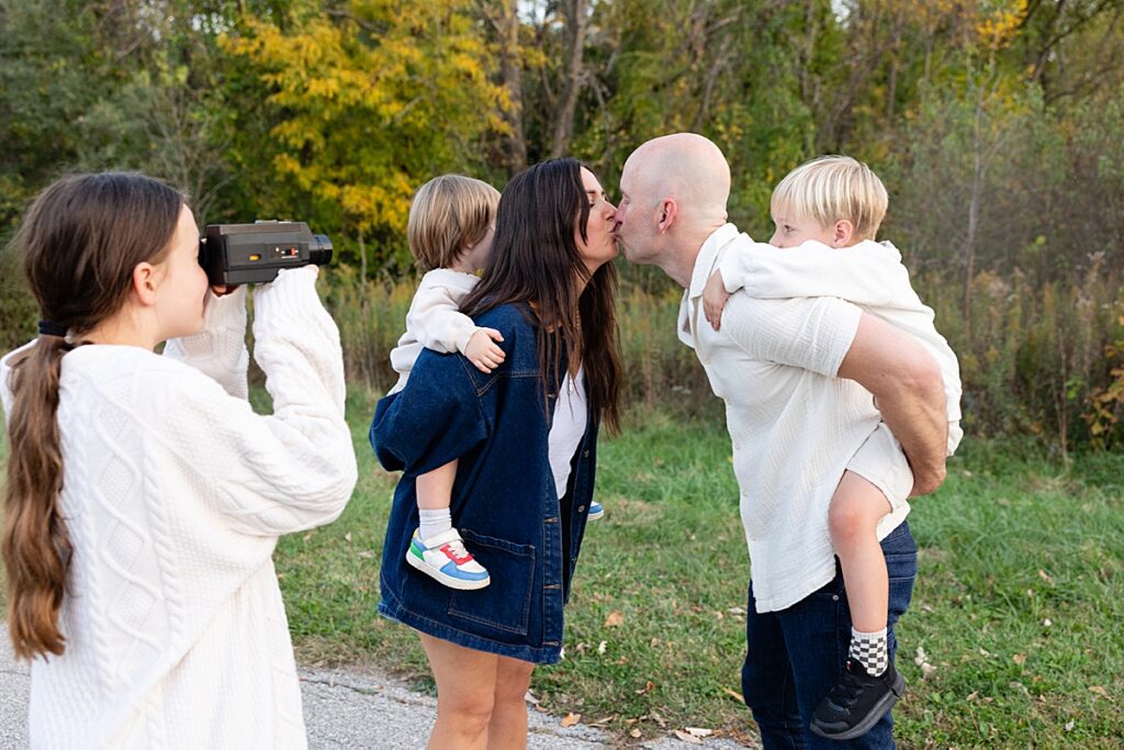 Young girl filming her parents kissing while they carry their kids piggyback, with trees and grass in the background.