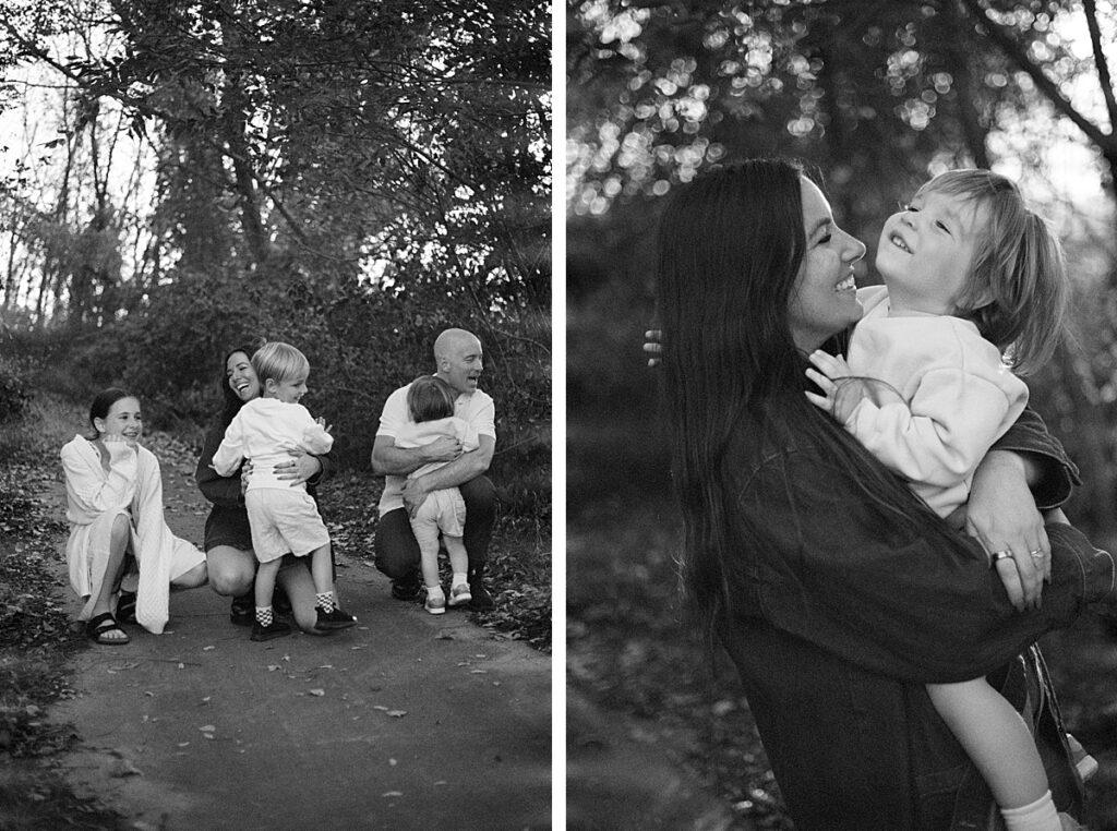 Family gathered on a wooded path, parents holding children in a natural moment captured in black and white. Mother holding her toddler son, smiling at each other during a fall session, in a softly lit black and white photo.