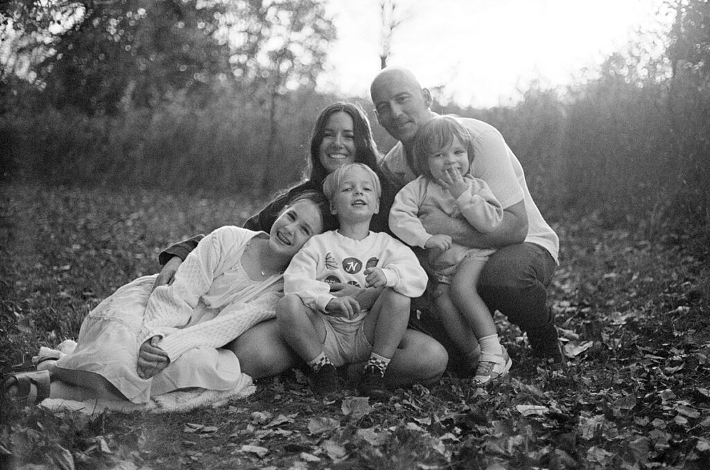 Family of five sitting close together on a blanket in a leaf-covered field, smiling in a timeless black and white photo