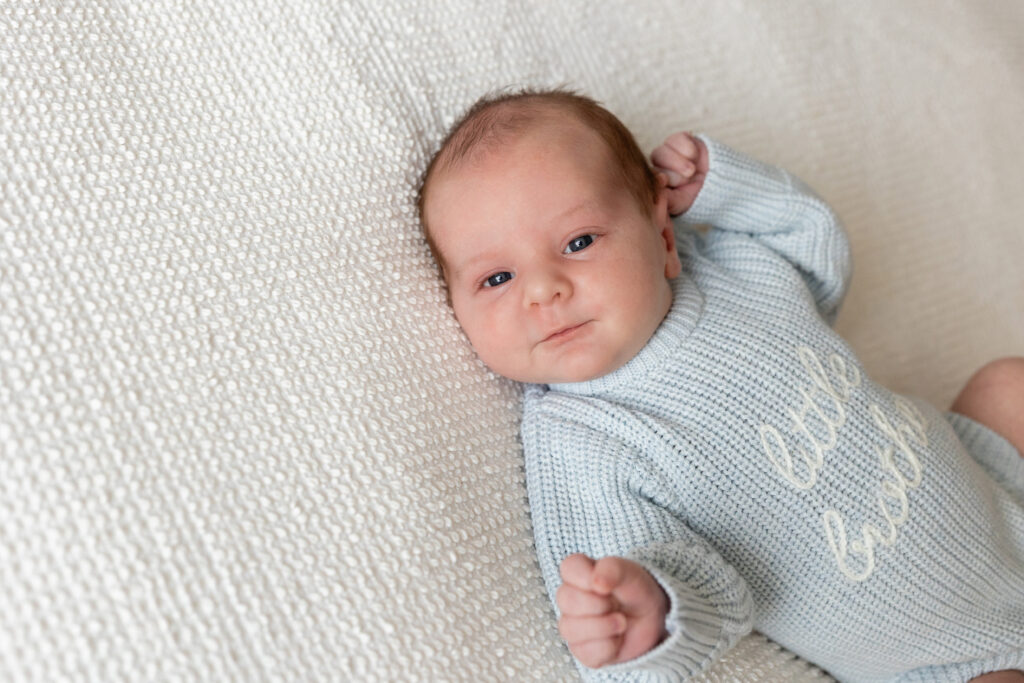 Newborn baby lying on a white textured blanket, wearing a light blue knit onesie with "little brother" stitched on the front.
