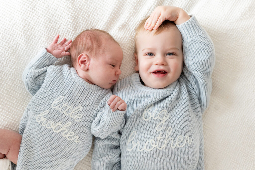 Toddler and newborn lying side by side on a bed, wearing matching knit sweaters labeled "big brother" and "little brother."