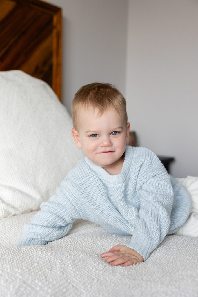 Toddler in a pale blue knit sweater crawling on a white bed, looking toward the camera with a slight smile.