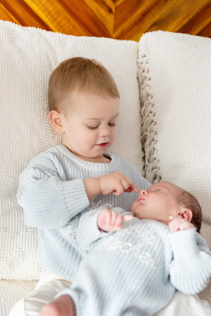 Toddler in a knit "big brother" sweater gently touching a newborn’s nose while sitting on a bed together.