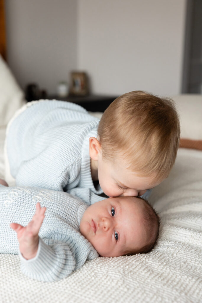 Toddler lying next to a newborn baby on a white bed, both wearing matching light blue knit "brother" sweaters.