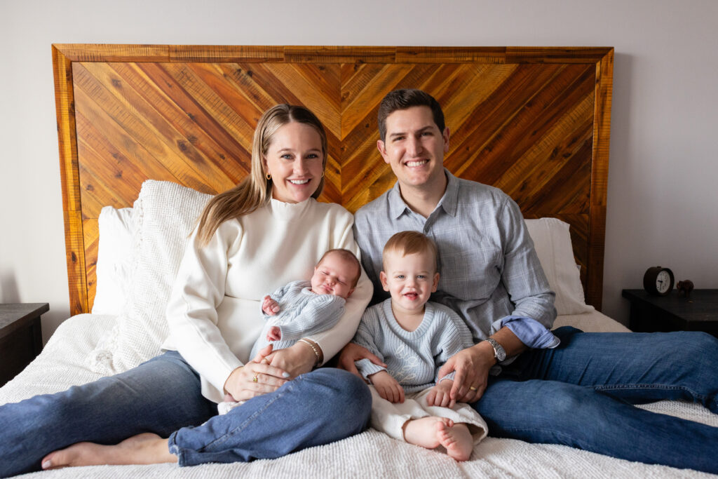Smiling family of four sitting on a bed, with parents holding their toddler and newborn dressed in matching knit outfits.