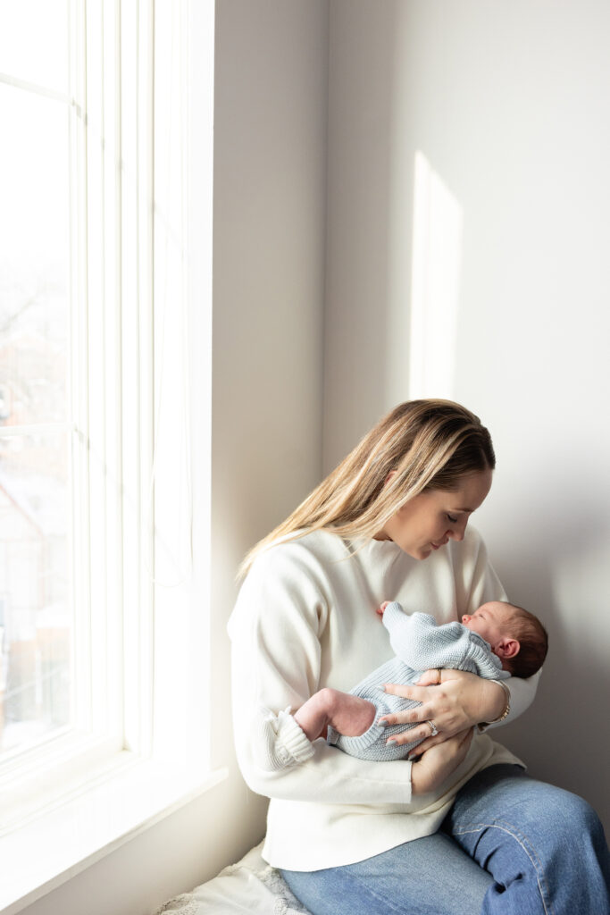Mother holding her newborn in a light-filled room, sitting by a large window in soft natural light.