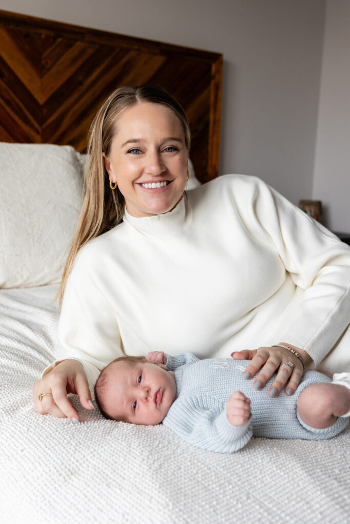 Mother lying on a bed beside her newborn in a light blue knit onesie, both resting on a white blanket.