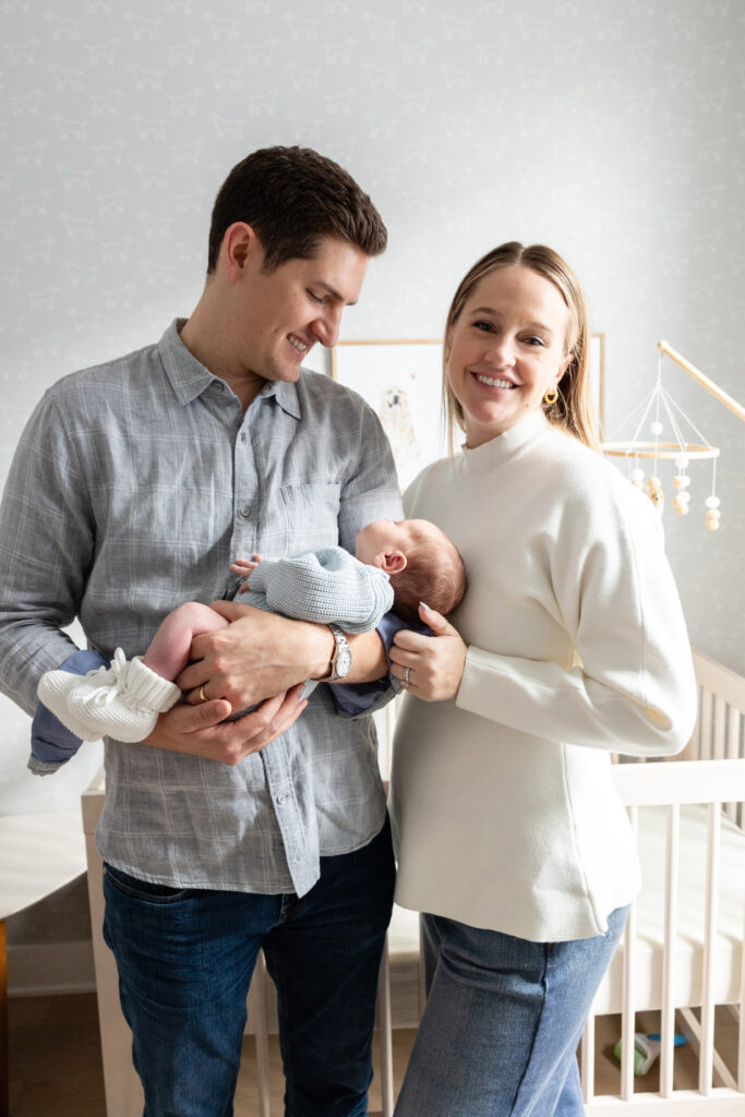 Mother and father smiling while holding their newborn together in a softly lit nursery with a crib in the background.
