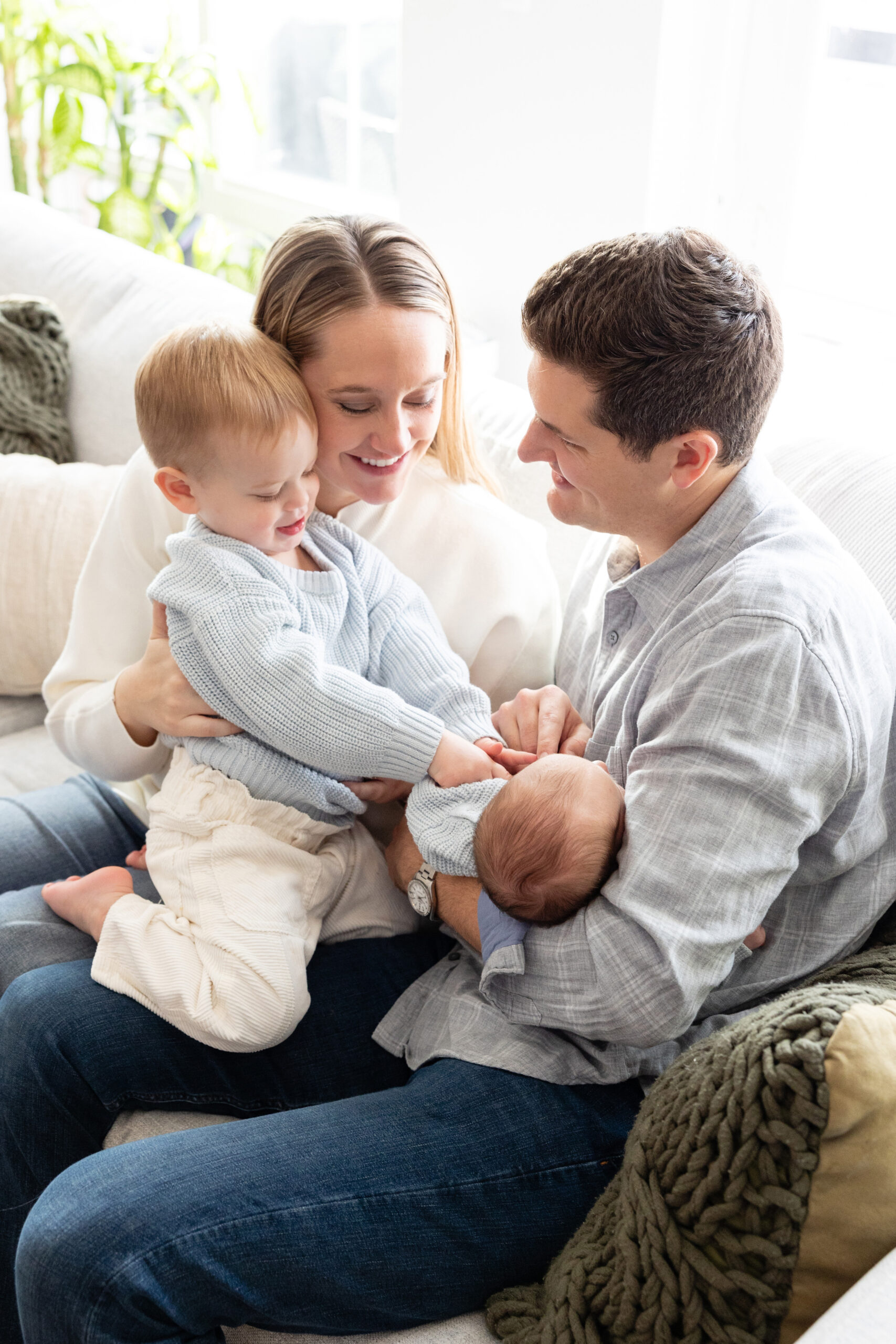Parents sitting on a couch with their toddler and newborn, all wearing cozy outfits in a light-filled living room.