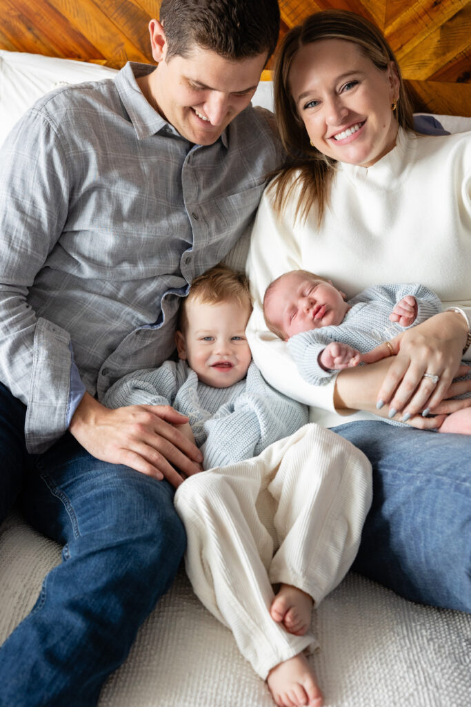 Close-up of parents sitting on a bed with their toddler and newborn, all dressed in soft knits and cozy textures.