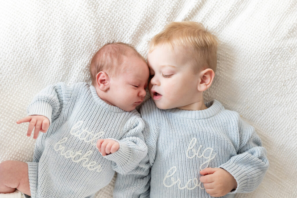 Newborn and toddler lying on a bed in matching “little brother” and “big brother” sweaters, heads gently touching.