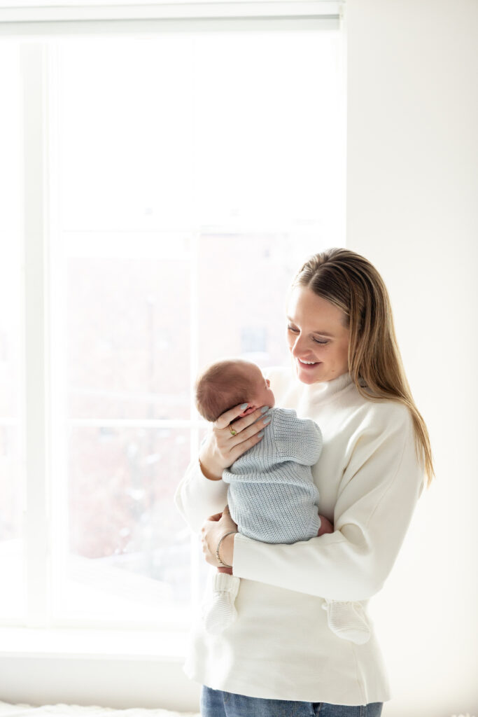 Mother standing in front of a large window, holding her newborn upright against her chest in natural light.