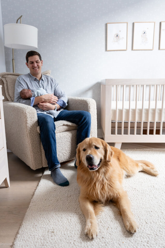 Father sitting in a cozy armchair holding a newborn, with a golden retriever lying on a white rug in a softly lit nursery.