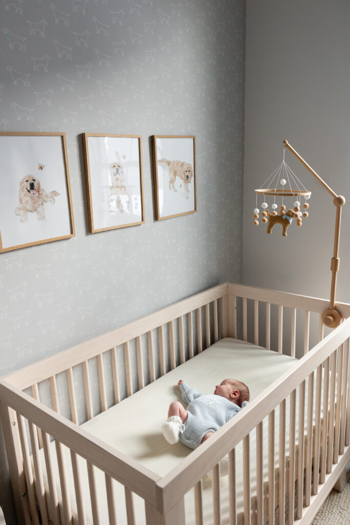Newborn lying in a light wood crib beneath framed dog illustrations and a wooden mobile in a dog-themed nursery.