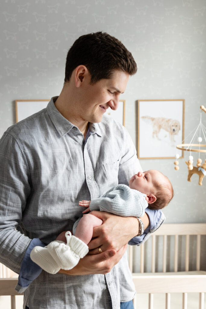 Father smiling while cradling a newborn in a light-filled nursery, with dog artwork and a mobile in the background.