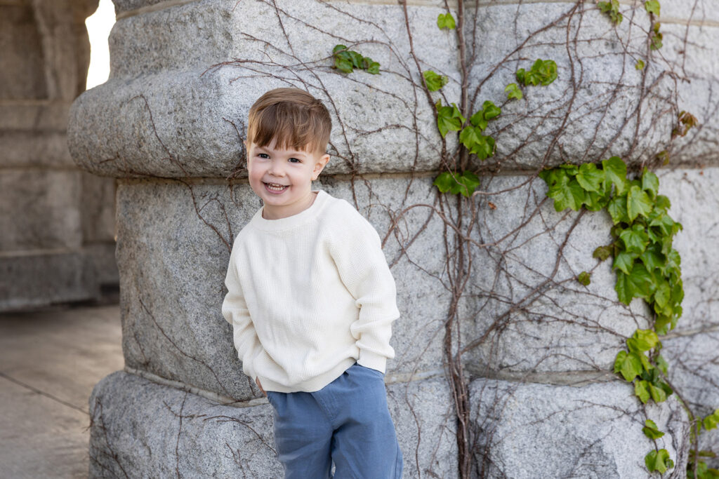 Young boy smiling beside an ivy-covered stone wall at Lincoln Park Nature Boardwalk.