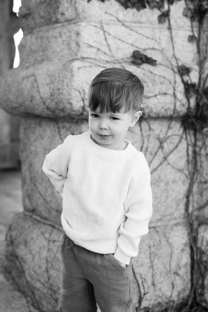Black and white portrait of a young boy posing beside an ivy-covered stone wall at Lincoln Park Nature Boardwalk.