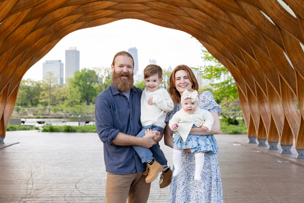 Parents holding their young children under the wood pavilion at Lincoln Park Nature Boardwalk with the Chicago skyline in the background
