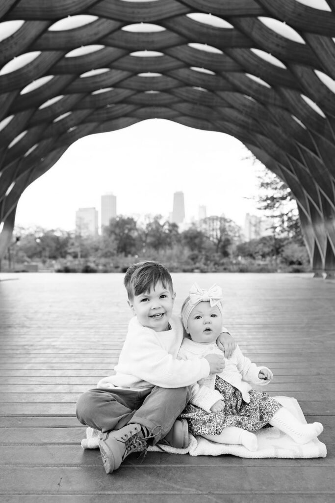 Black and white portrait of brother hugging his baby sister while sitting on a blanket beneath the pavilion at Lincoln Park Nature Boardwalk