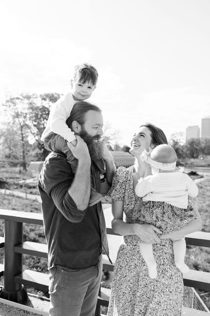 Black and white photo of young boy sitting on father's shoulders and mother holding baby on the bridge at Lincoln Park Nature Boardwalk in Chicago.