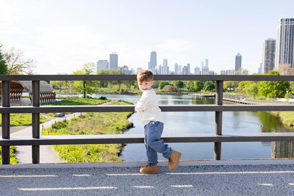 Young boy smiling on a bridge overlooking the Lincoln Park Nature Boardwalk with the Chicago skyline behind him.